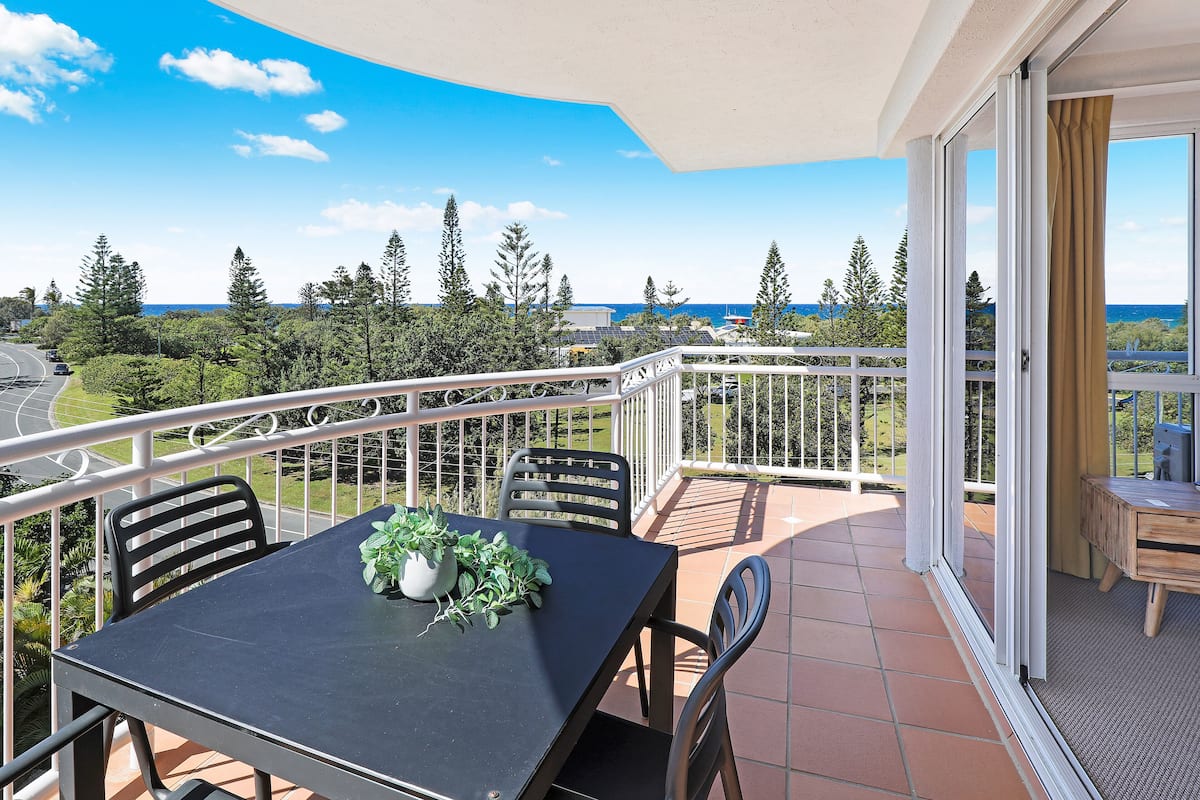 A balcony with a table and chairs, a potted plant, and a view of the ocean and trees.