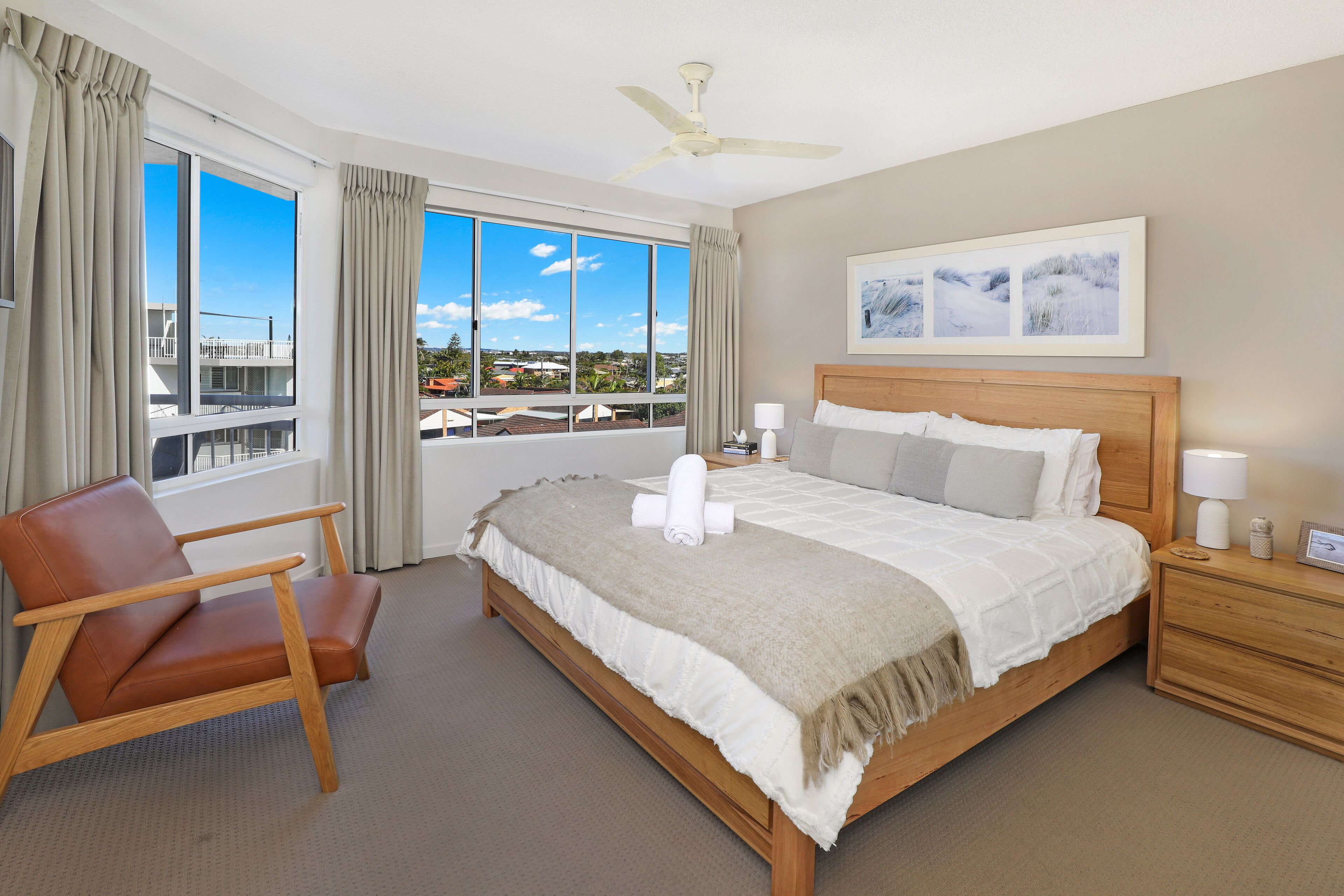 A hotel room with a large bed, a wooden nightstand, a chair, and a view of the city through large windows.