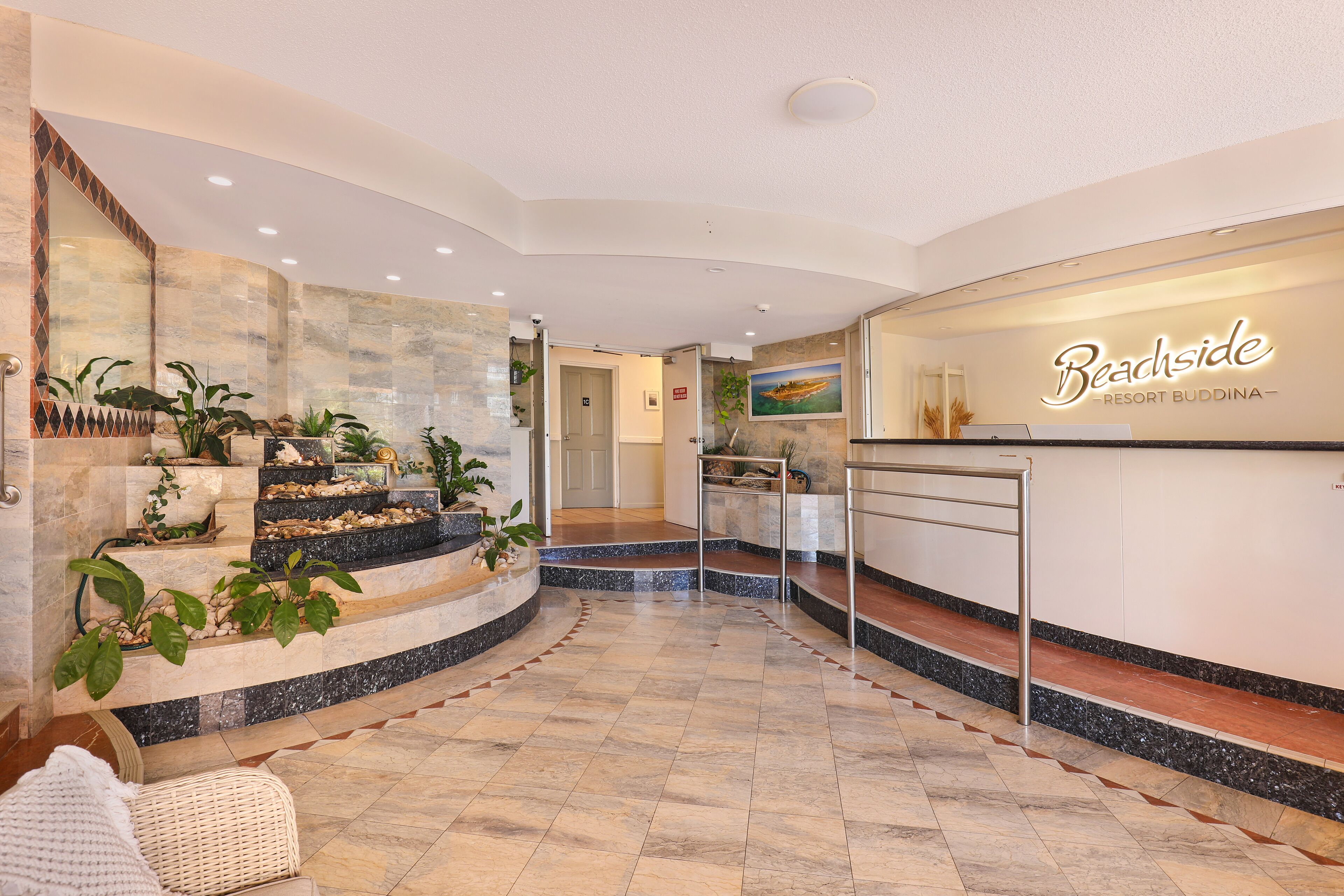 Un moderno vestíbulo de hotel con una escalera curva, un mostrador de recepción y diversas plantas.