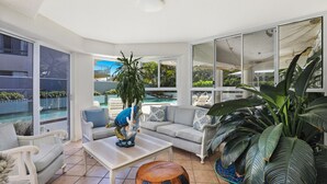 A sunlit living room with a view of a pool, a sofa set, a coffee table, and large green plants.