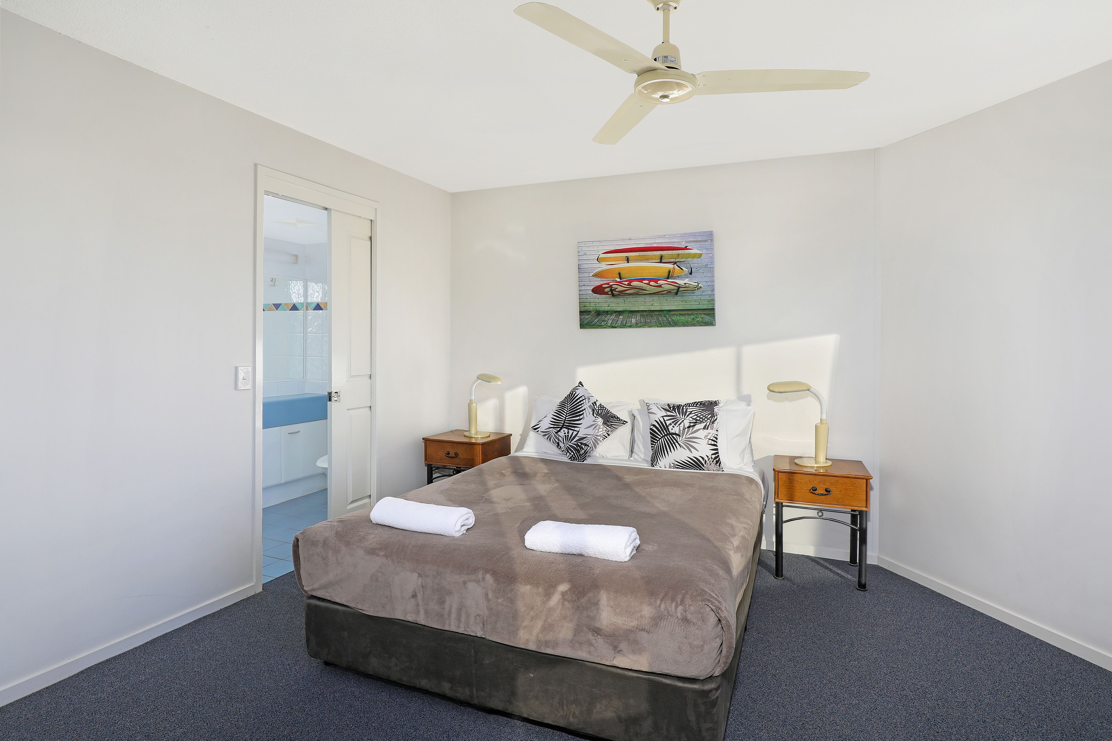 A bedroom with a bed, two bedside tables, a ceiling fan, and a painting on the wall.