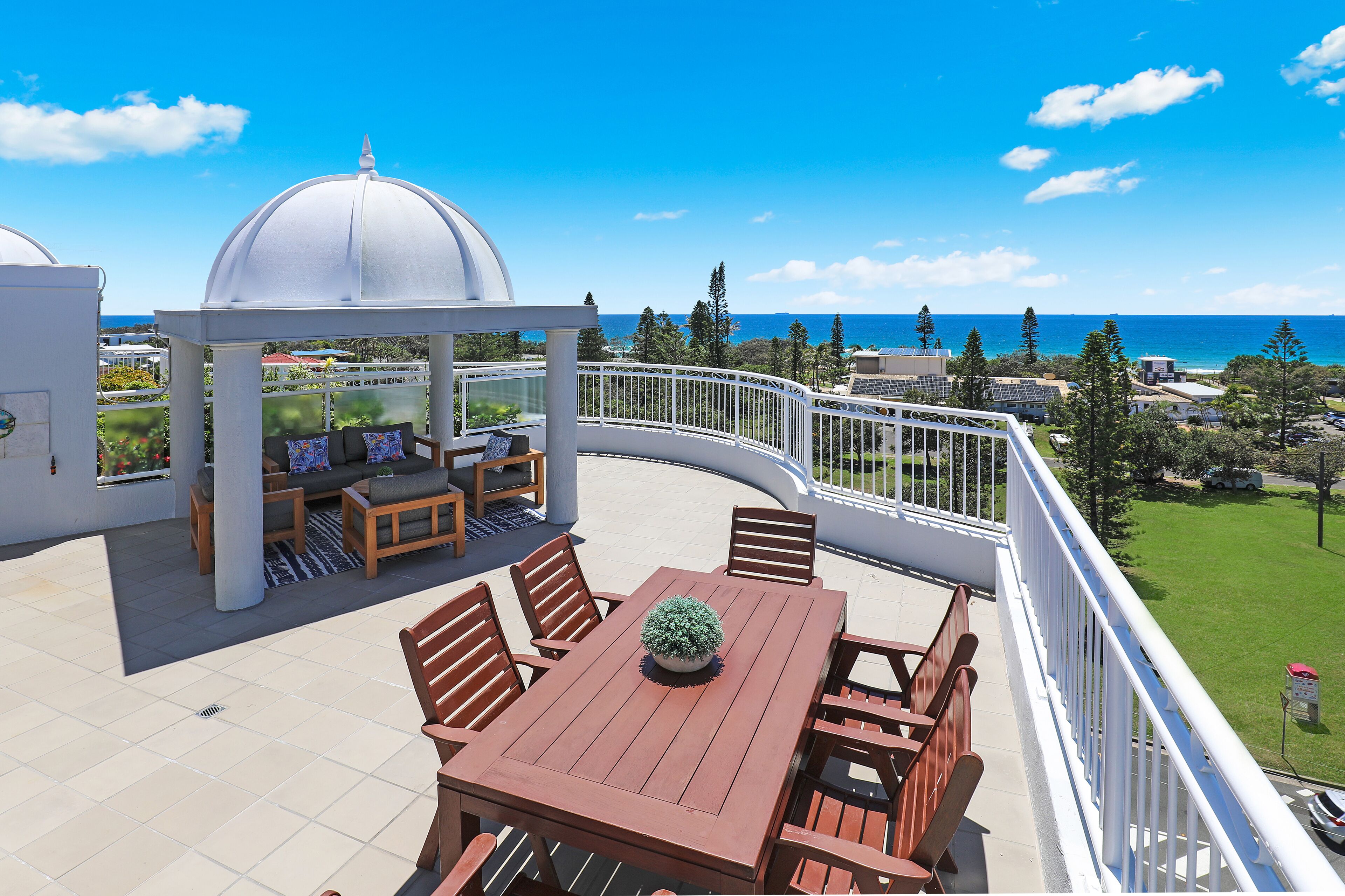 A rooftop terrace with a wooden table and chairs, a white gazebo, and a view of the ocean.
