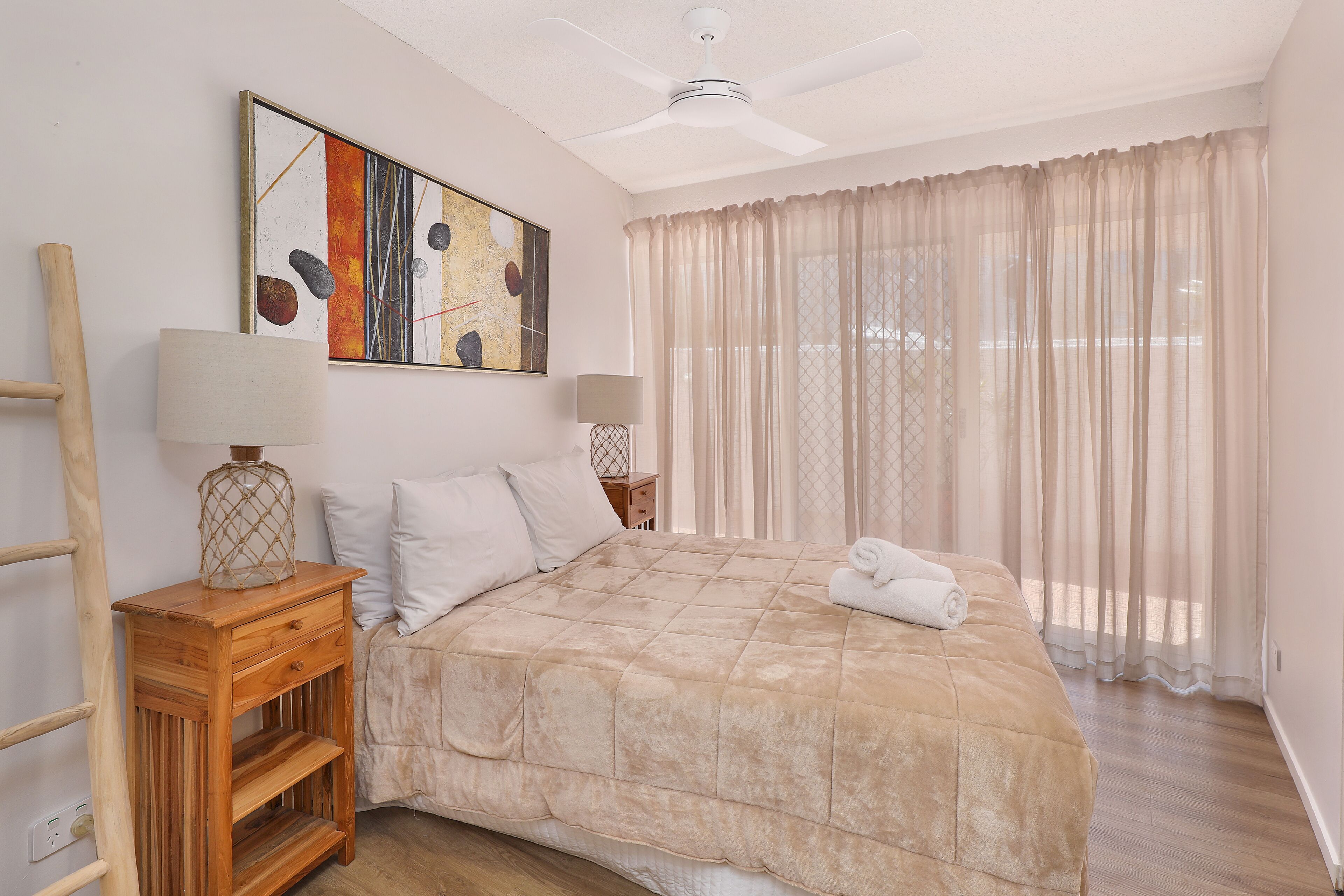 Un dormitorio con una cama, dos mesitas de noche, un ventilador de techo, una ventana con cortinas traslúcidas y un cuadro colgado en la pared.