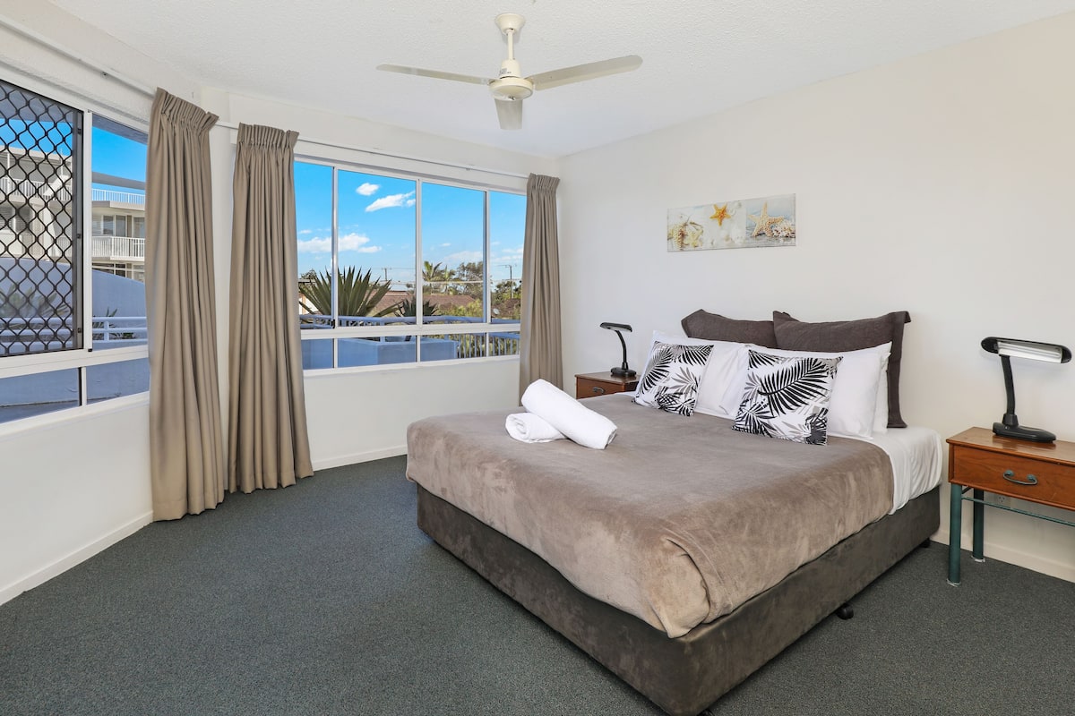 A hotel room with a large bed, a ceiling fan, a nightstand, a lamp, and a window with curtains.