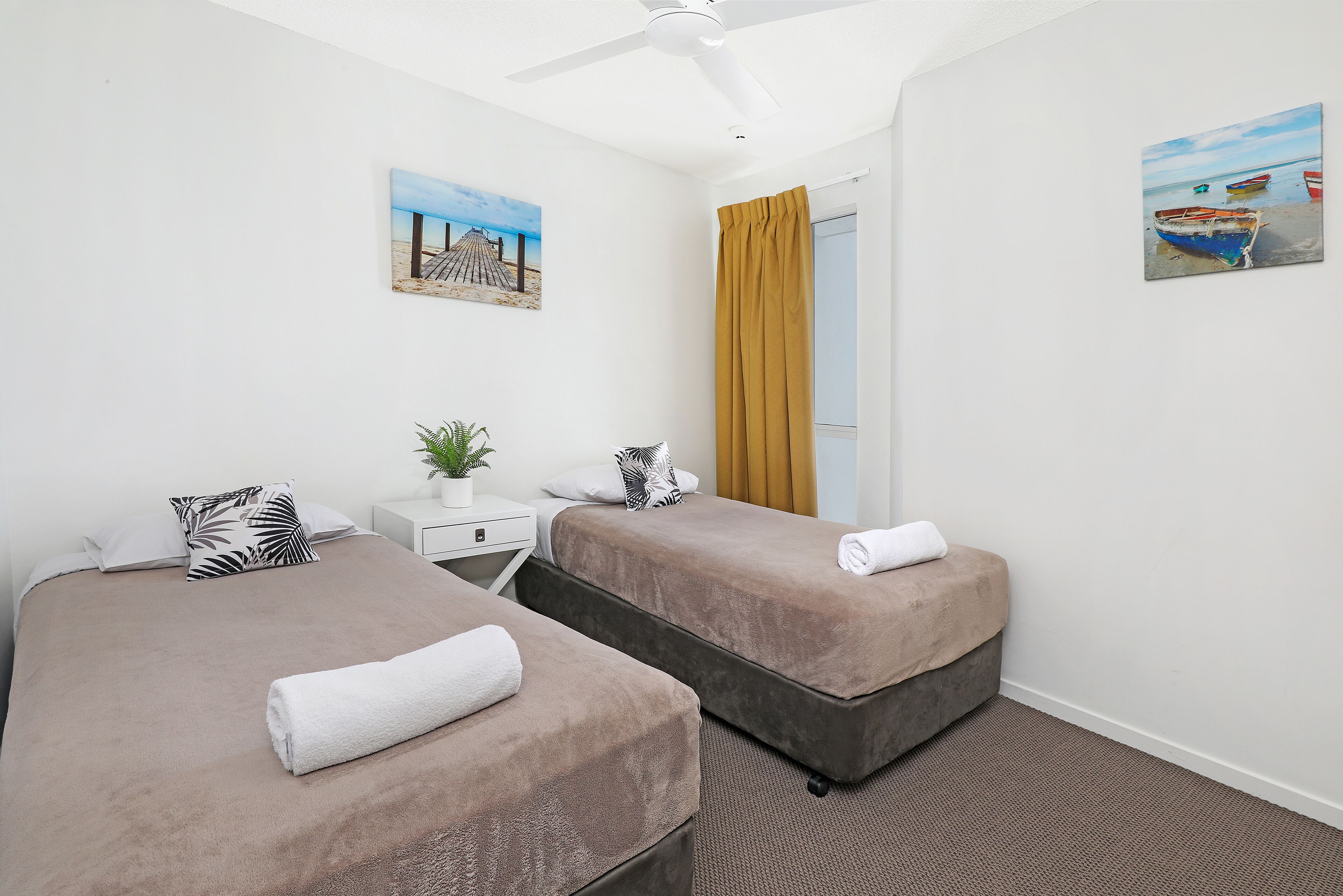 A hotel room with two beds, a ceiling fan, a small table with a plant, and two paintings on the walls.