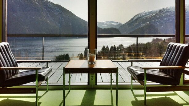 Une pièce avec vue sur un lac et des montagnes, deux chaises, une table et un verre d’eau.