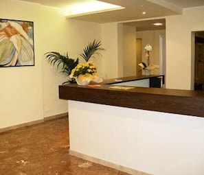 Un mostrador de recepción con encimera de madera oscura y base blanca, una planta en maceta con flores amarillas y una obra de arte enmarcada en la pared.