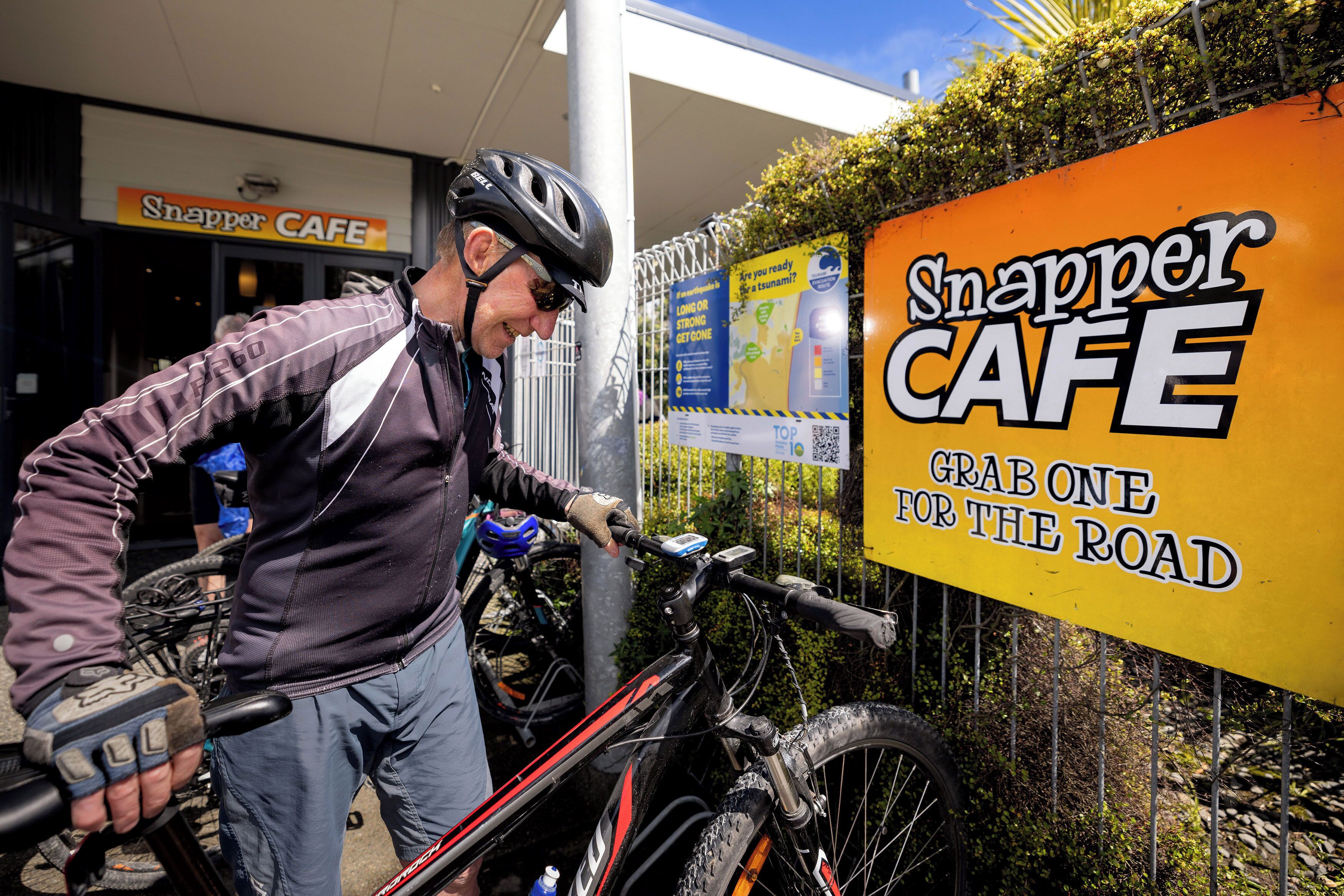 Eine Person steht neben einem Fahrrad vor einem Gebäude, an dem ein Schild mit der Aufschrift 'Snapper Cafe' hängt.