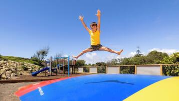 A child jumping on a blue and yellow trampoline with a playground in the background.
