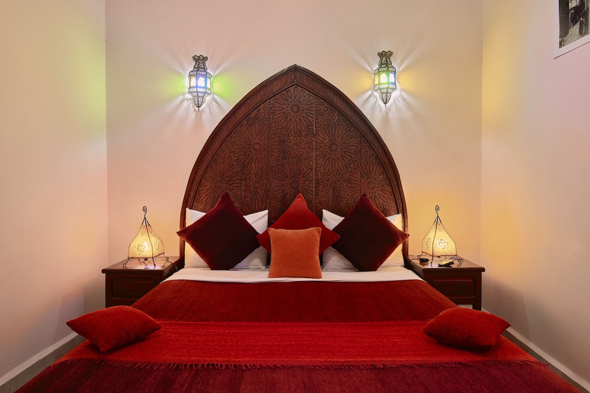 A bed with a red bedspread and multiple pillows, a wooden headboard with intricate carvings, and two wall-mounted lamps.