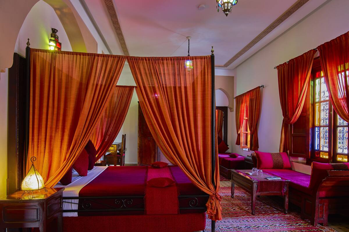 A room with a bed, a sofa, and a table. There are curtains and a chandelier.