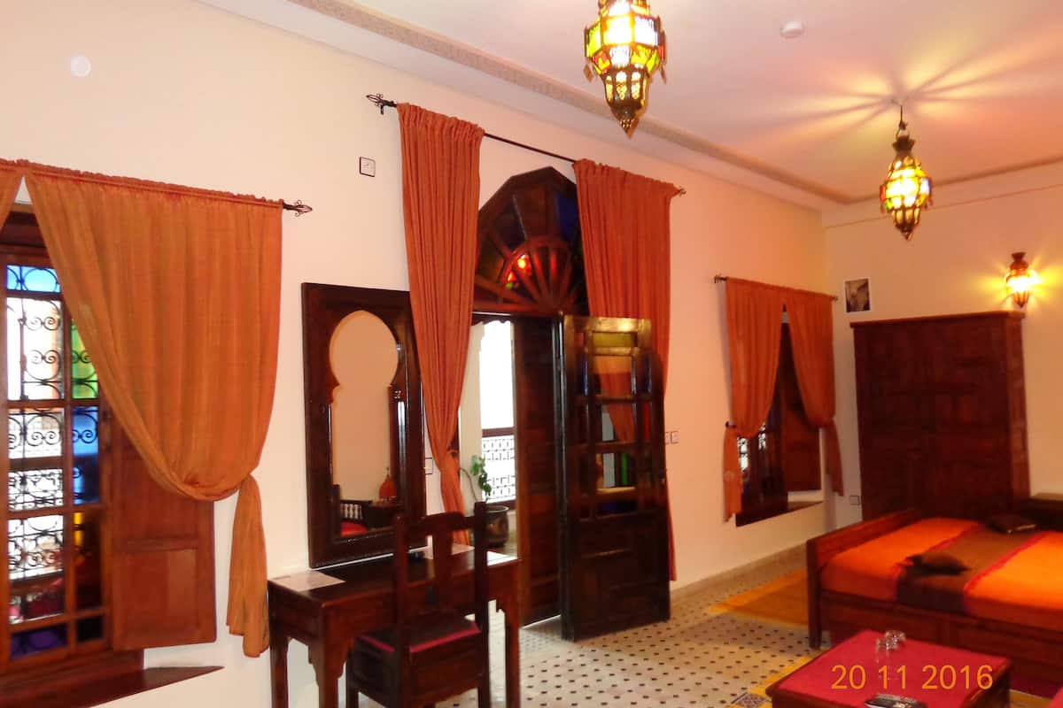A room with a bed, a wooden chair, a small table, and a mirror. There are curtains on the windows and a chandelier hanging from the ceiling.