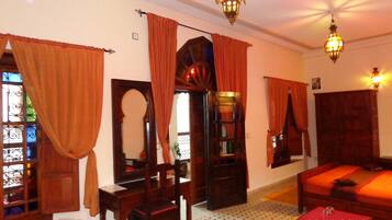 Una habitación con una cama, una silla de madera, una mesita y un espejo. Hay cortinas en las ventanas y un candelabro colgando del techo.