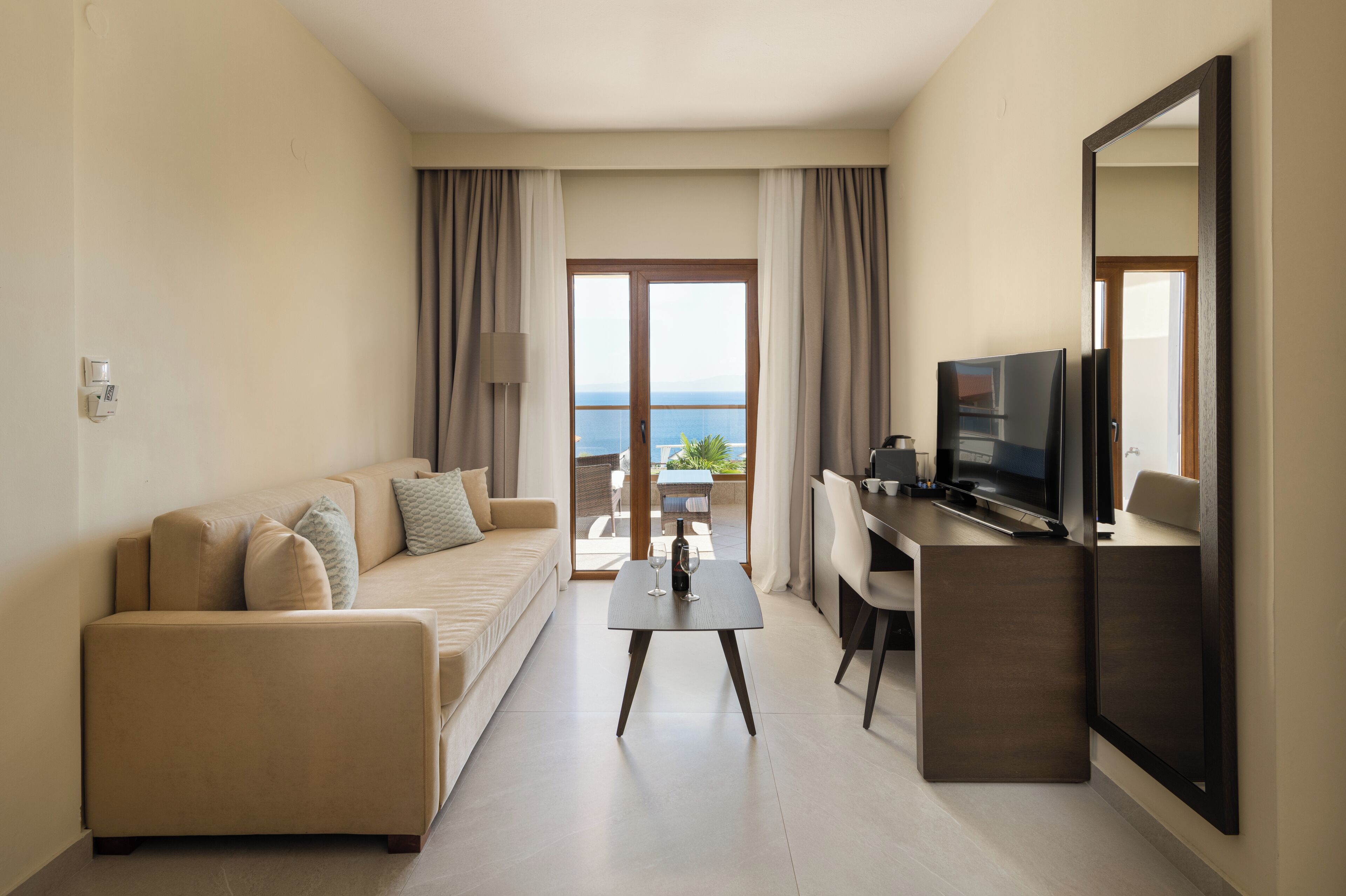 Quarto de hotel moderno com sofá, uma pequena mesa com uma taça de vinho, televisão e varanda com vista para o mar.