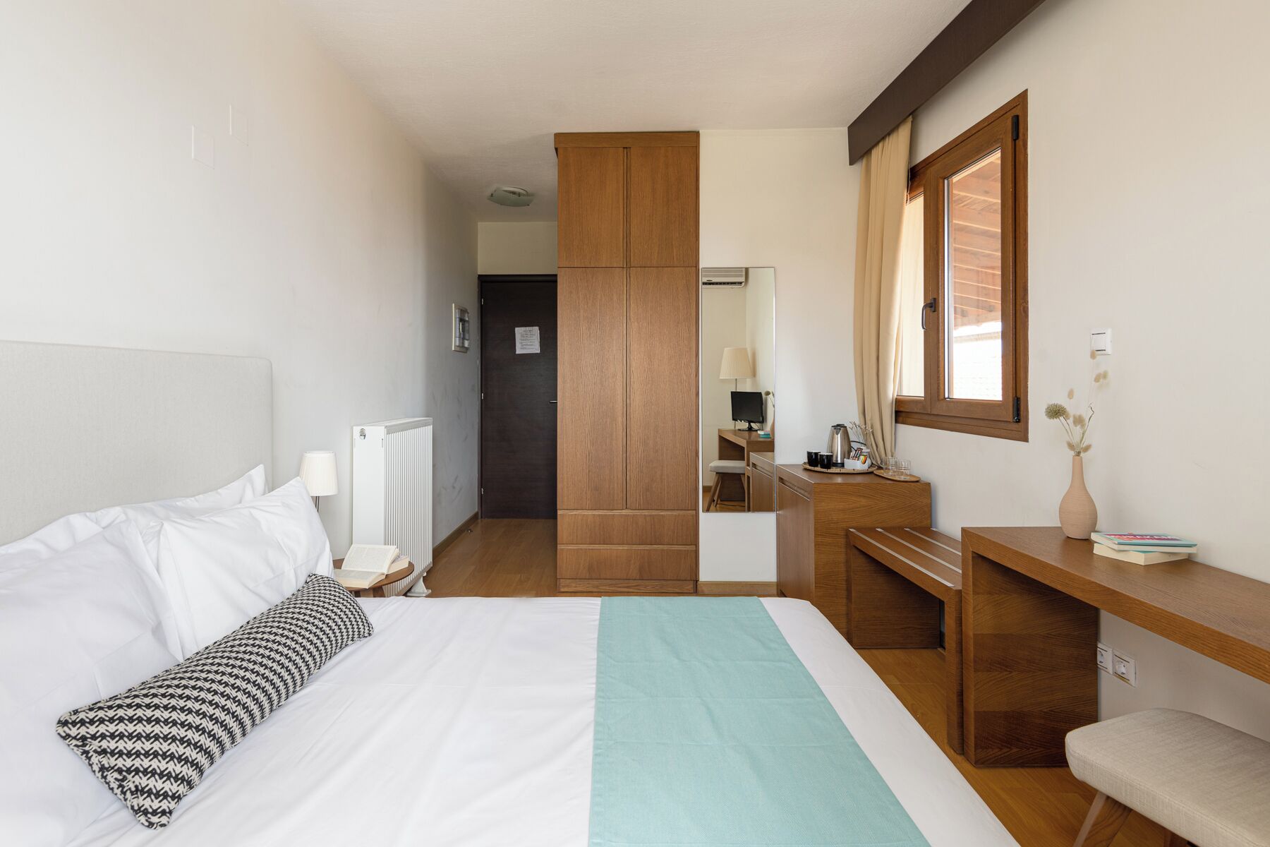 A hotel room with a bed, a desk, a chair, a radiator, a mirror, and a window.