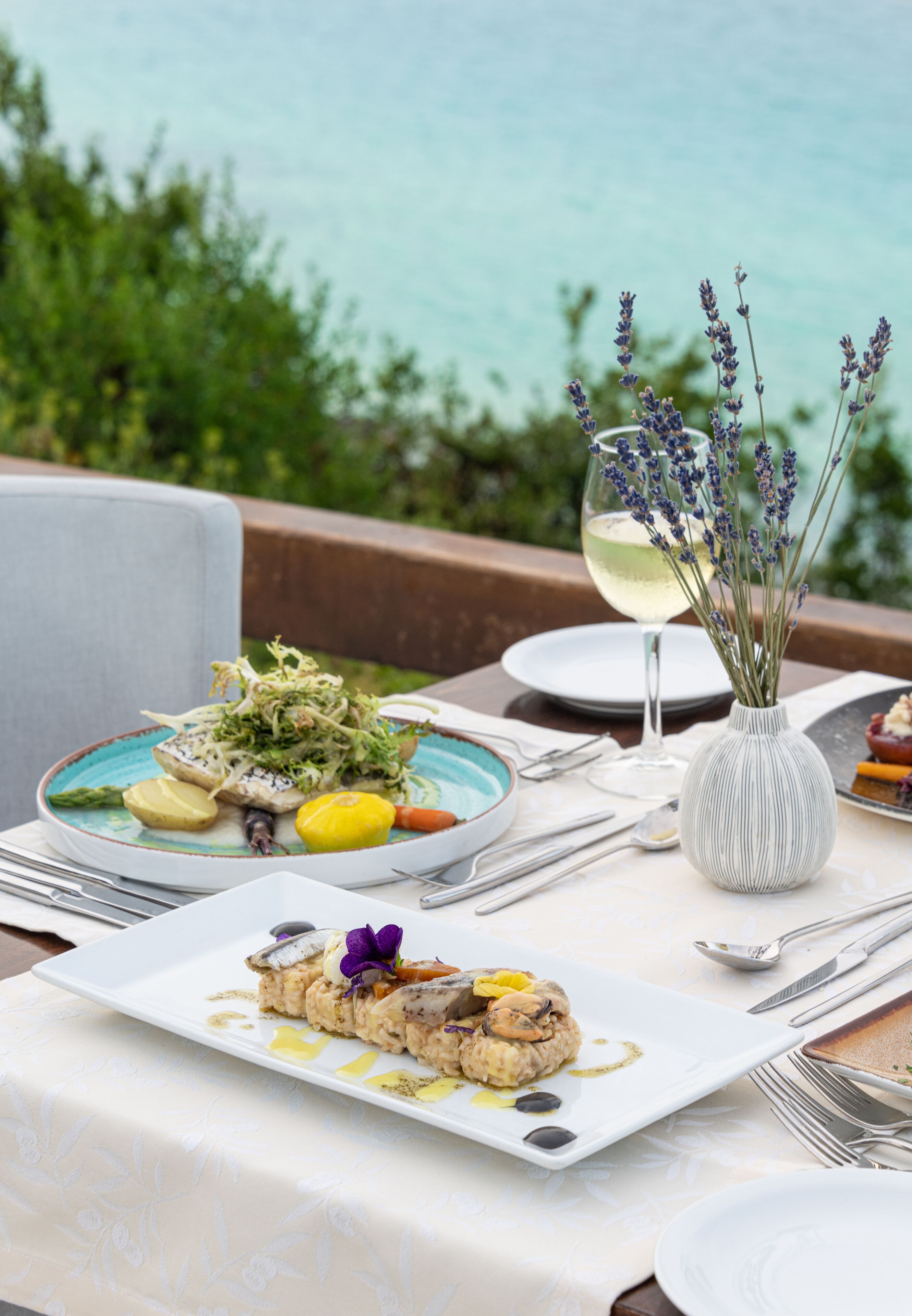 A table set with plates of food, a glass of white wine, and a vase with lavender.