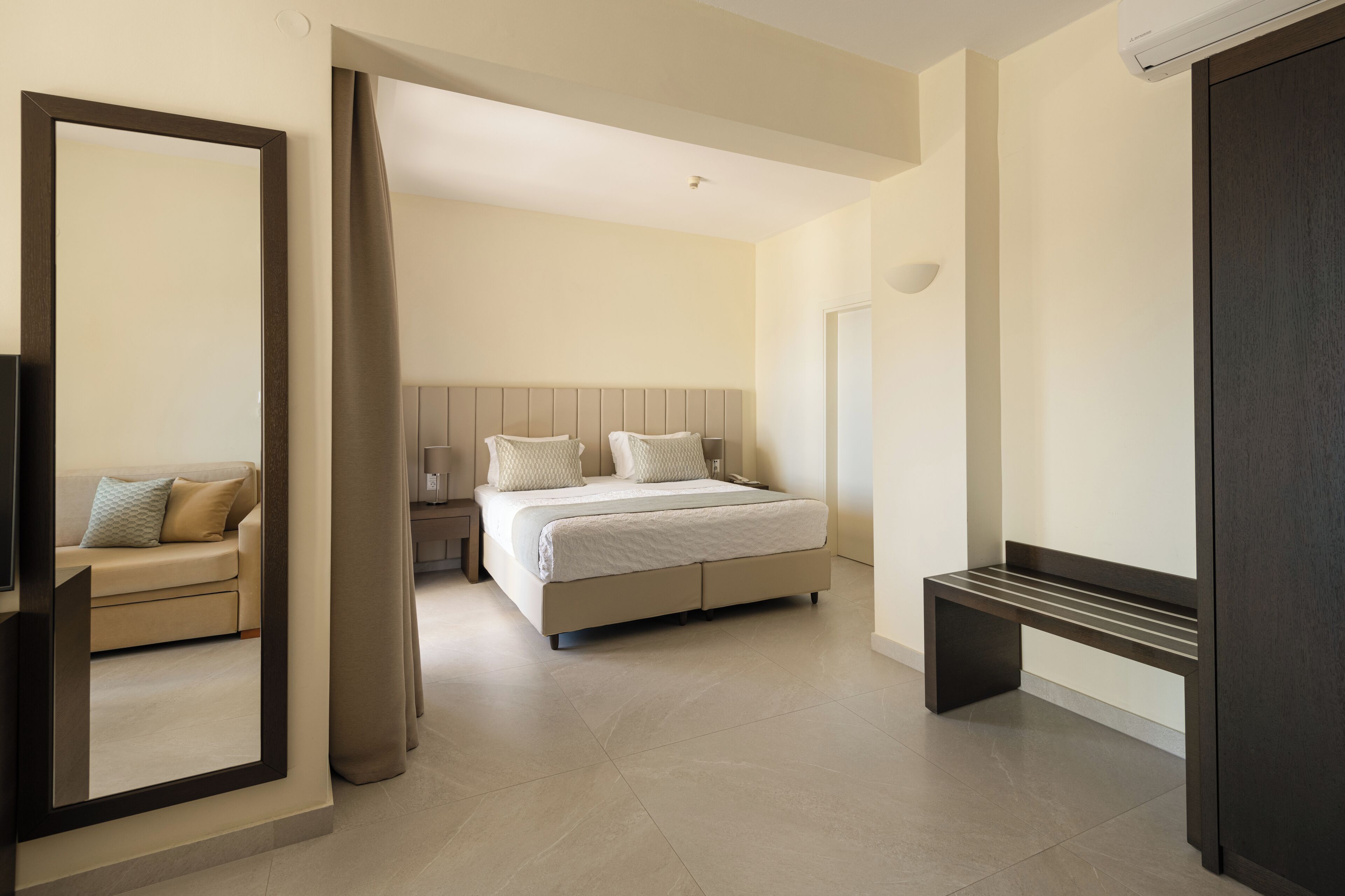 A modern hotel room with a large bed, a bedside table, a bench, and a mirror.
