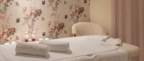 A neatly made bed with white linens and towels, a chair, and a wall with floral wallpaper.