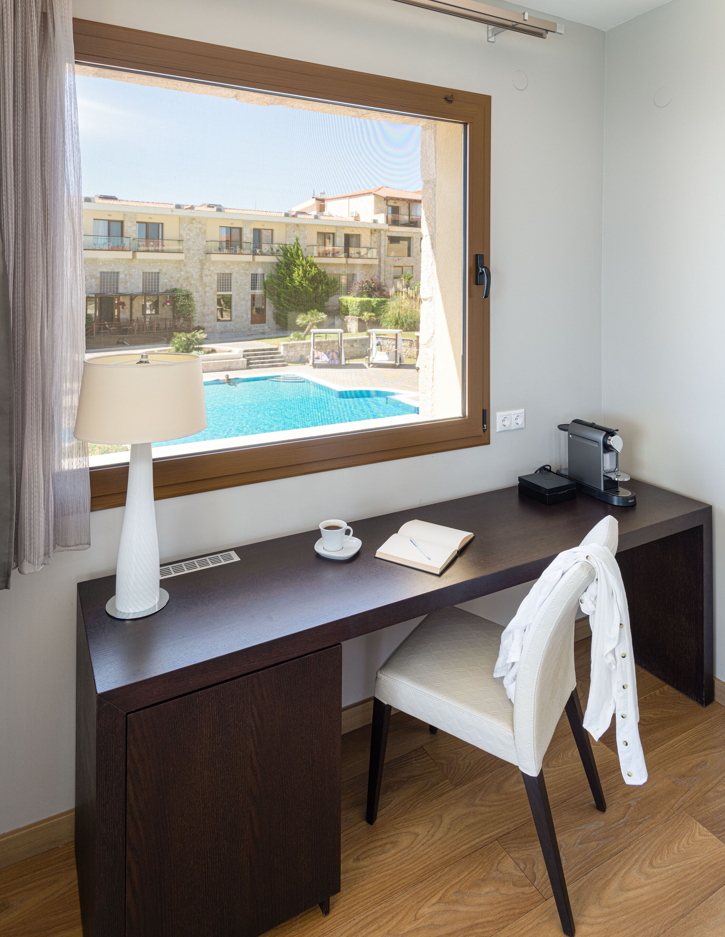 Una habitación con escritorio, silla, lámpara, taza de café, libreta y vistas a una piscina y edificios.