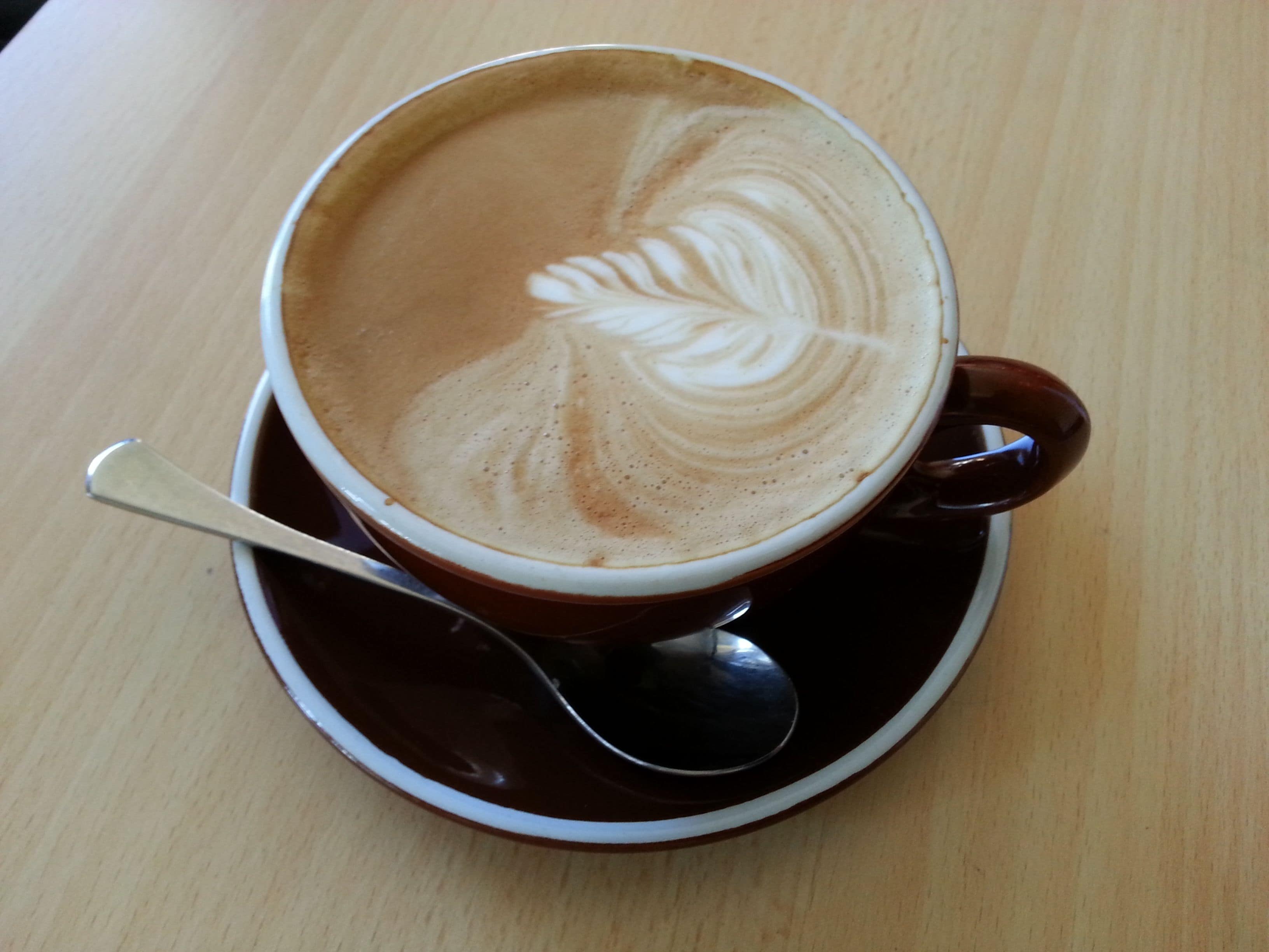 A cup of coffee with latte art on a saucer and spoon on a wooden table.