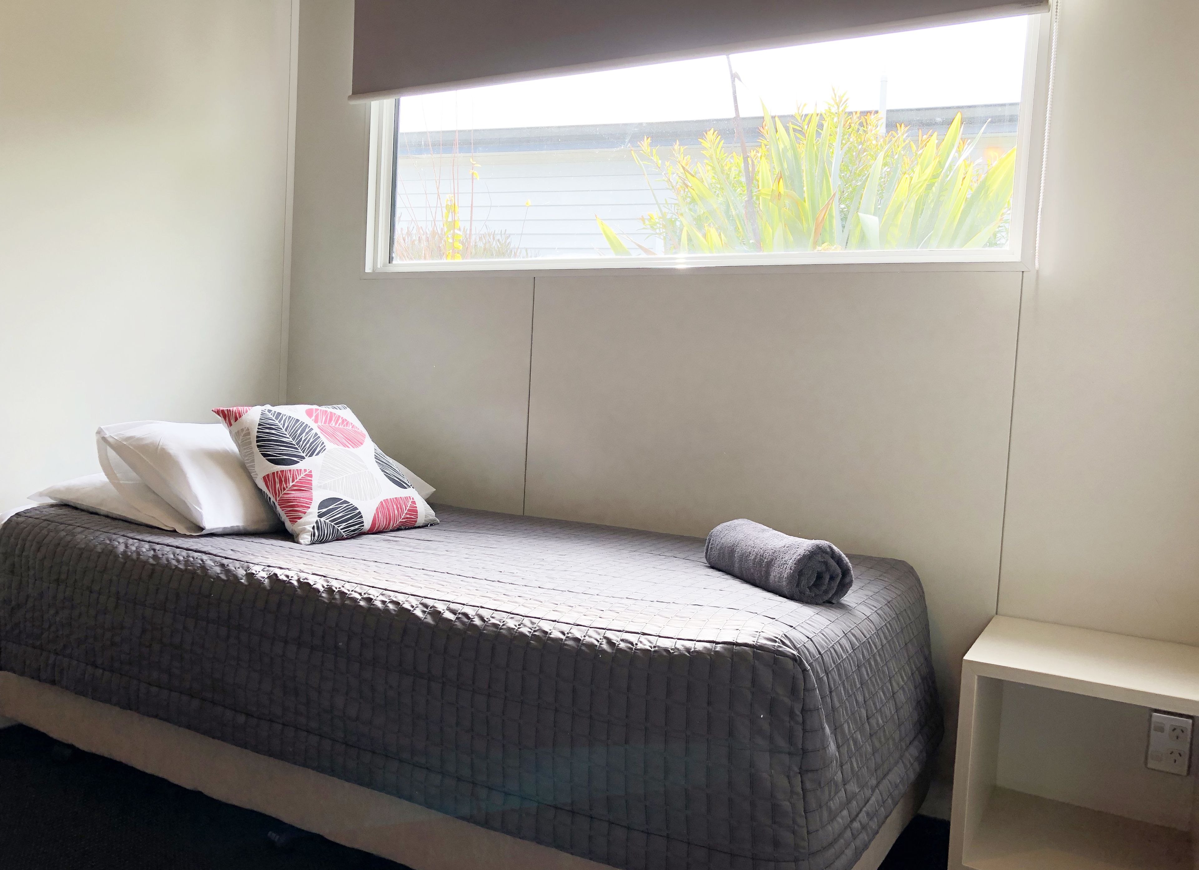 A bed with a grey quilt, two pillows with a leaf pattern, and a rolled towel. A window with a view of greenery and a building is above the bed.