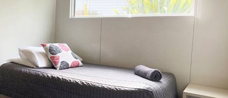 A bed with a grey quilt, two pillows with a leaf pattern, and a rolled towel. A window with a view of greenery and a building is above the bed.