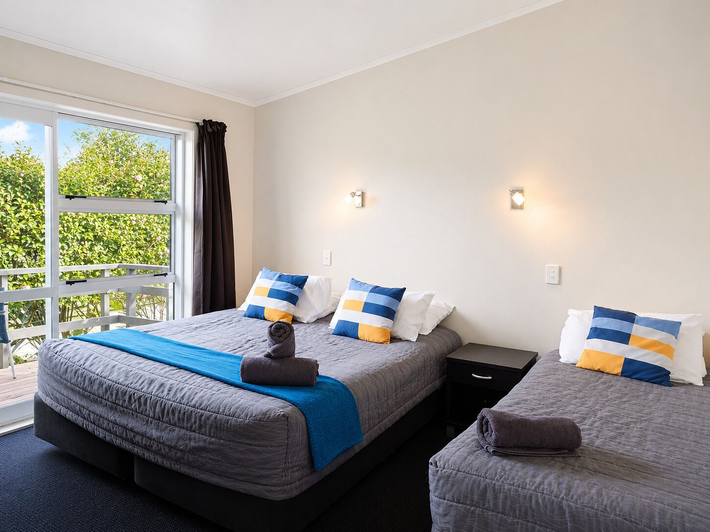 A hotel room with two beds, a large window, and a view of greenery outside.