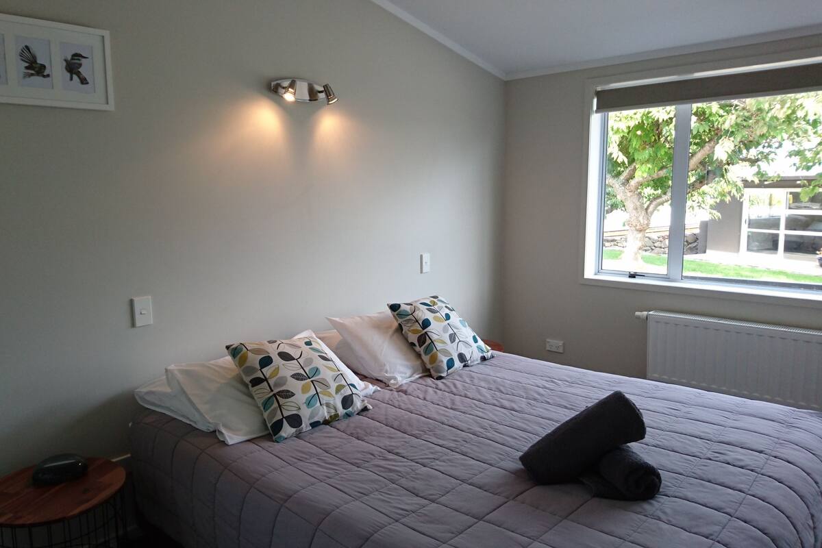 A bedroom with a bed, pillows, a bedside table, a wall-mounted light fixture, and a window with a view of greenery outside.