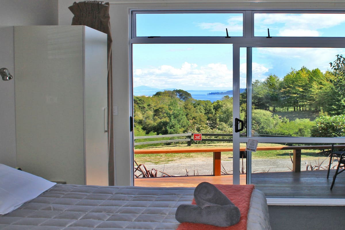 A bedroom with a bed, a view of a forest and a body of water through a large window.