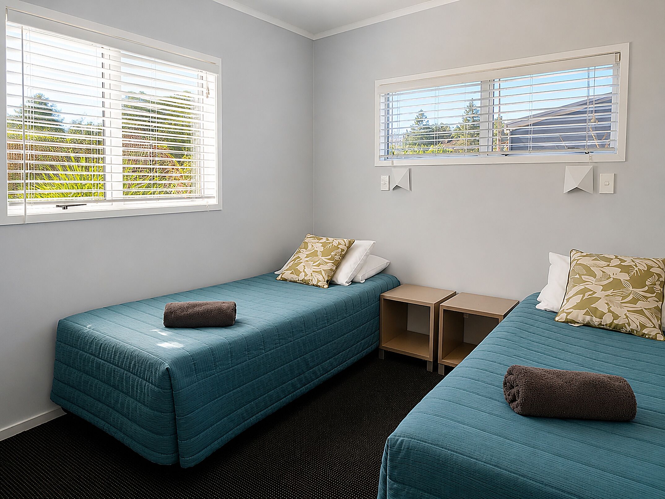 A room with two single beds, each with a pillow and a rolled towel, a small wooden nightstand, and a window with blinds.