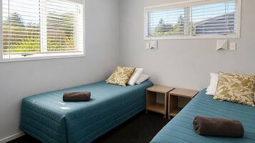 A room with two single beds, each with a pillow and a rolled towel, a small wooden nightstand, and a window with blinds.
