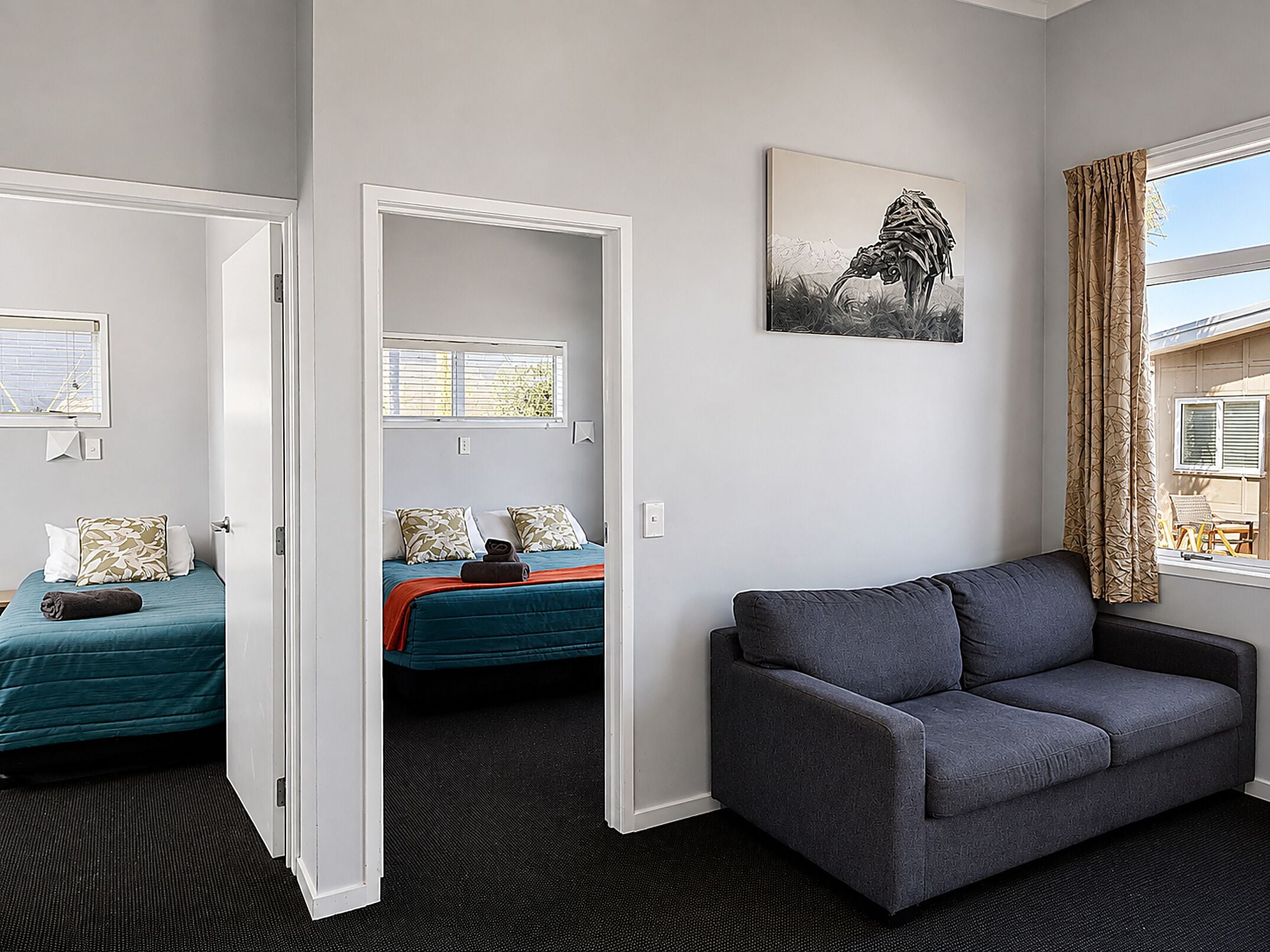 Una habitación de hotel con un sofá gris, una cama con ropa de cama color turquesa y un cuadro en la pared.