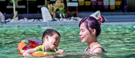 Ein Schwimmbecken mit klarem Wasser, ein Kind auf einem bunten Schwimmsack und eine Frau mit lila Haaren.
