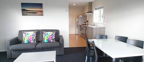 A modern living room with a grey sofa, colorful cushions, a white coffee table, and a dining table with chairs. A kitchen is visible in the background with a window and a view of the outside.