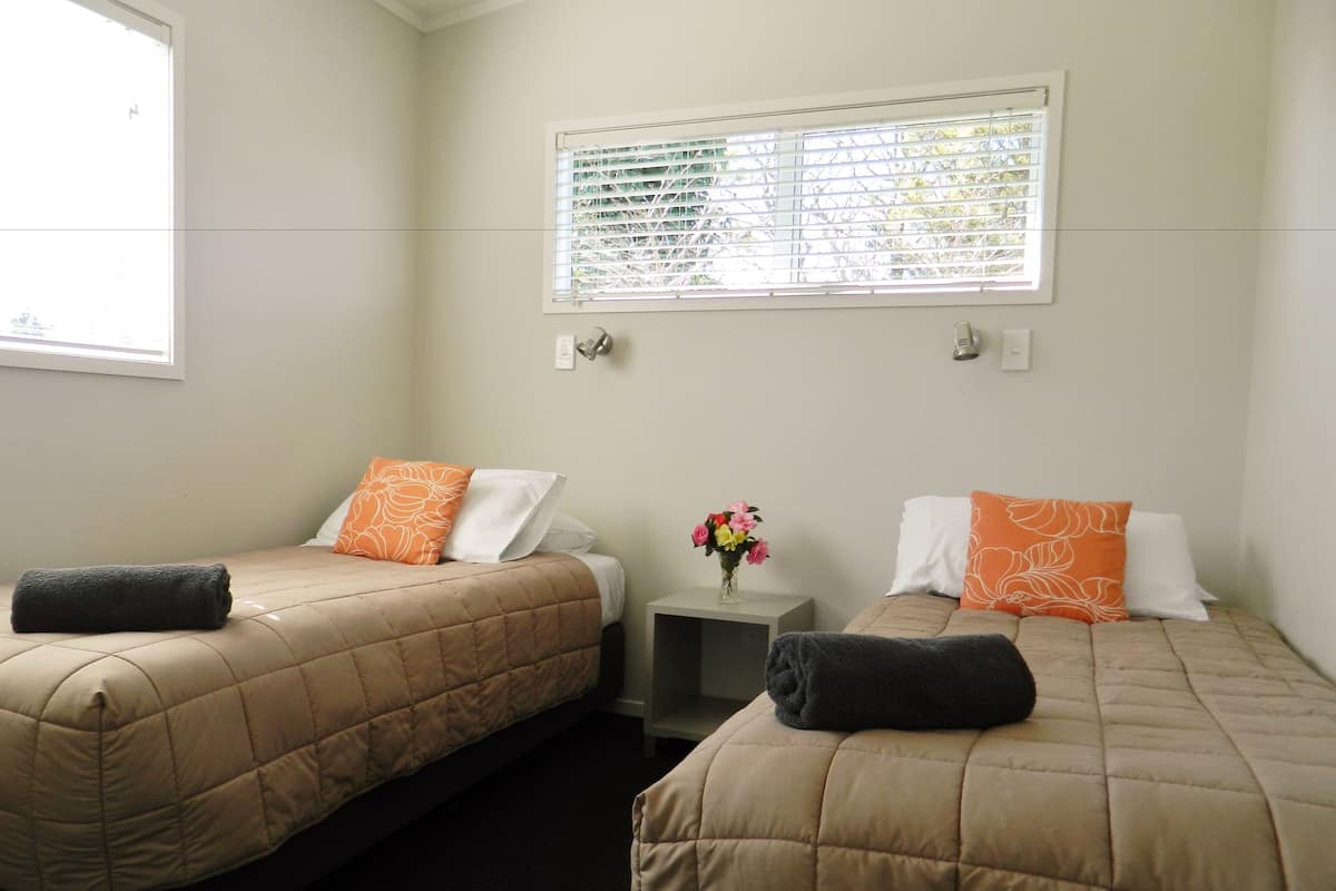 A hotel room with two beds, each with a beige quilted bedspread and matching pillows. There is a small table with a vase of flowers between the beds.