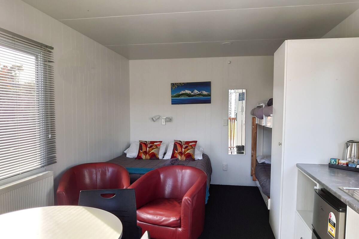 A compact hotel room with a bed, a small kitchenette, a dining table, and a red leather chair.