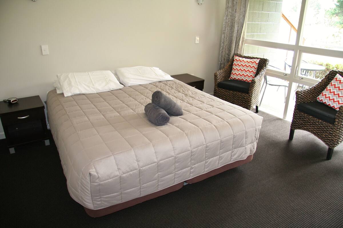 A neatly made bed with a beige quilt, two pillows, and a pair of folded towels on the bed. There are two wicker chairs with patterned cushions and a small table in the background.
