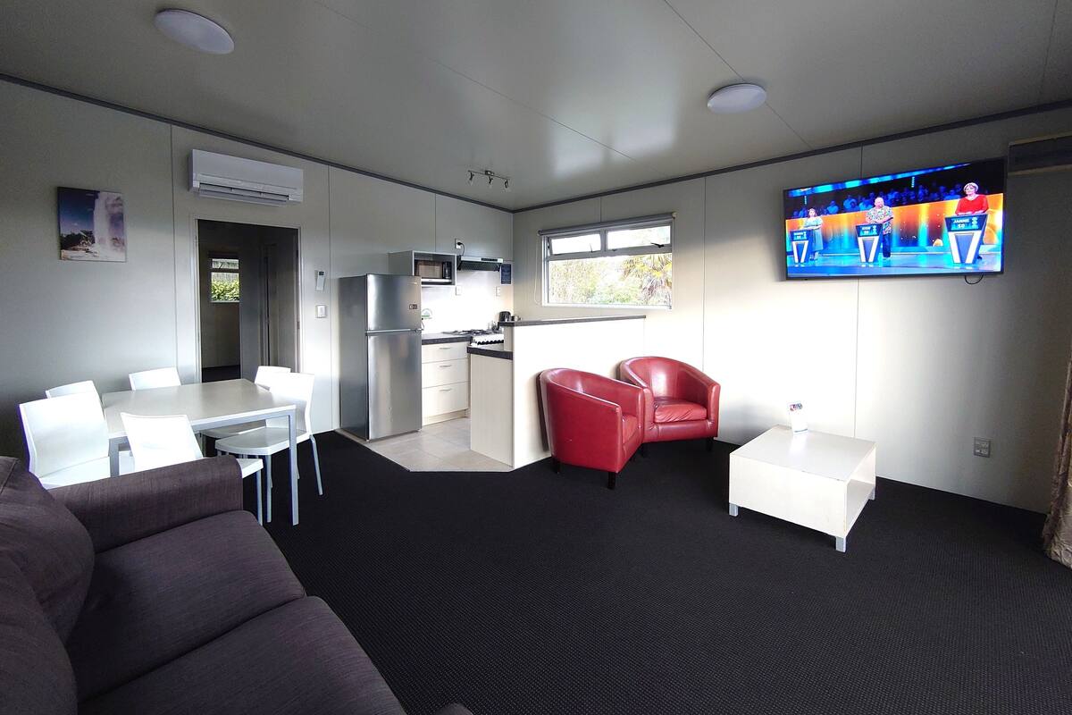 A modern living room with a sofa, a dining table, a kitchen area, and a TV mounted on the wall.