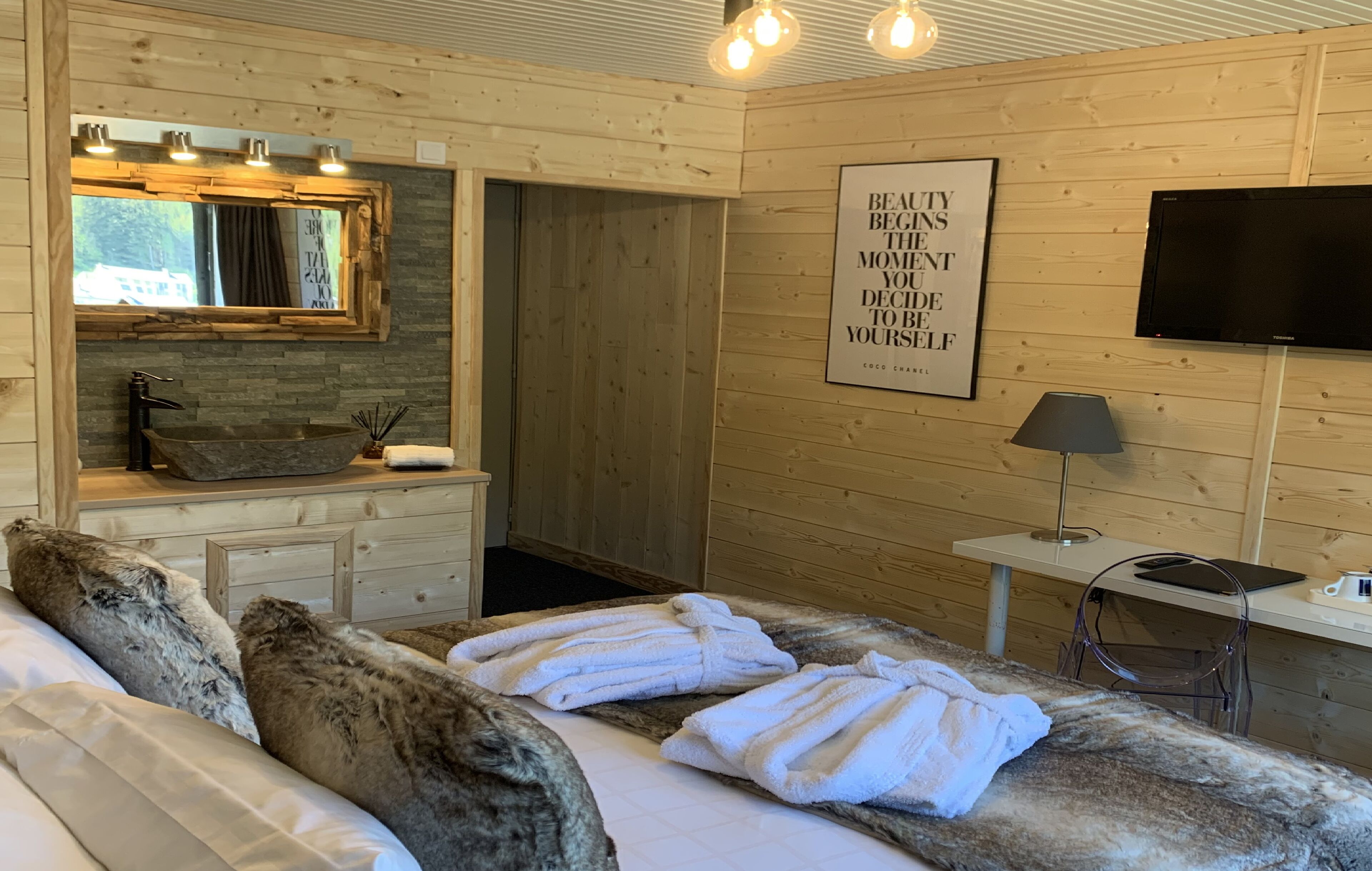 Ein gemütlicher Holzraum mit einem Bett, einem Schminktisch mit Spiegel, einem Fernseher und einem motivierenden Zitat an der Wand.