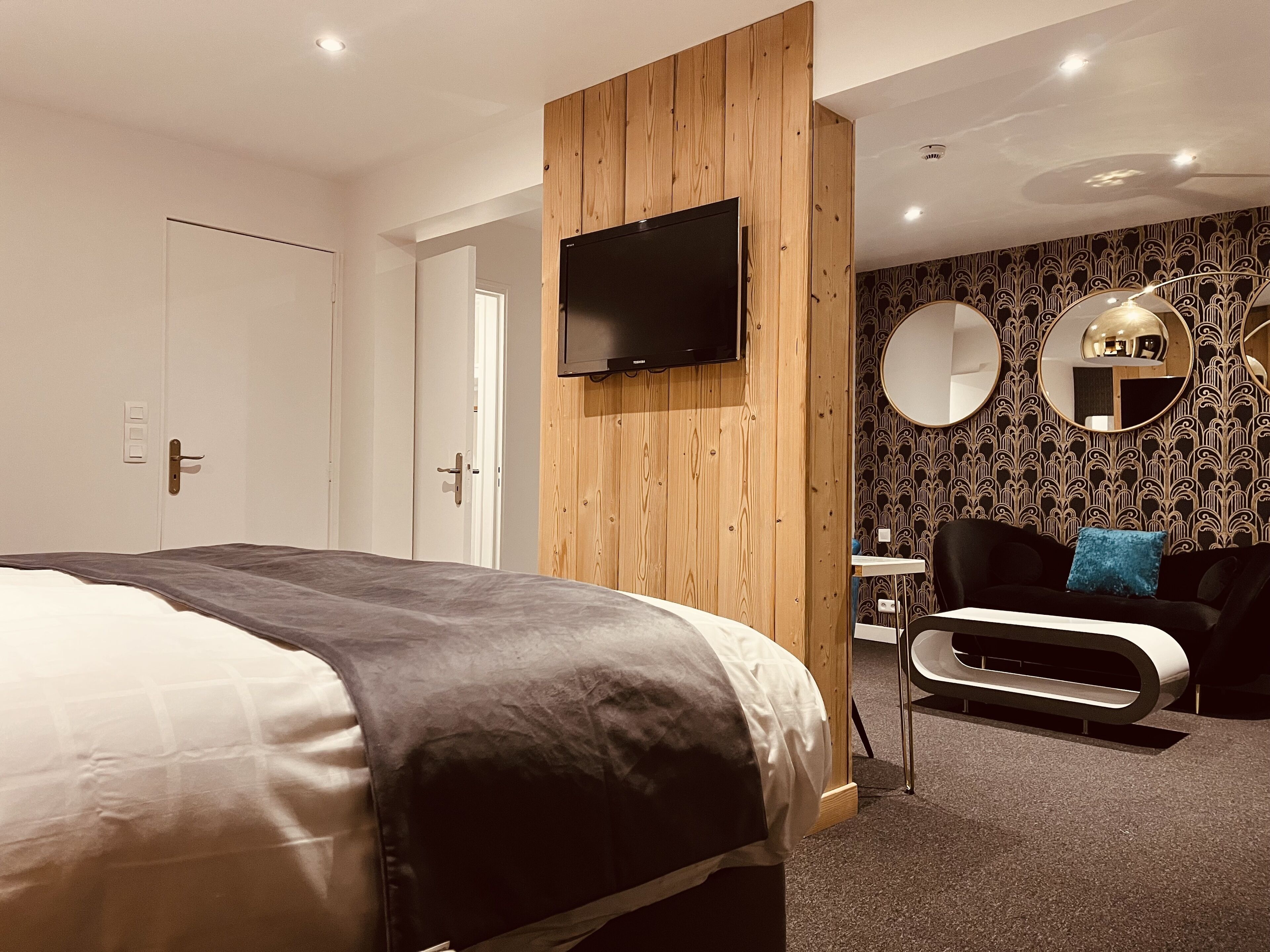 Une chambre d’hôtel moderne avec un lit, un téléviseur fixé sur un panneau en bois, un canapé noir avec un coussin bleu et une petite table avec un miroir.