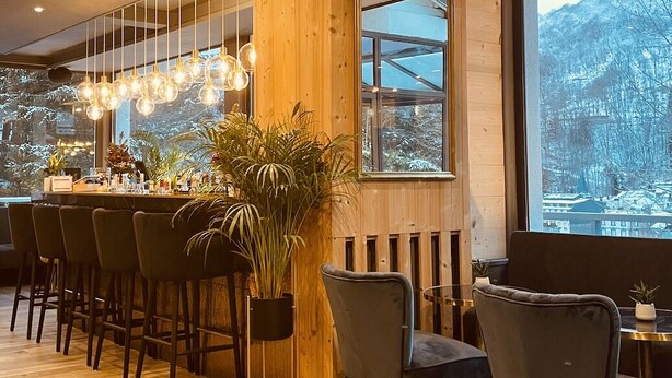 A modern bar area with wooden accents, a large window with a view of snow-covered trees, and hanging light fixtures. -  The Originals Boutique, Hôtel Le Panorama, Mont-Dore (Mont-Dore)
