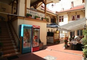 A narrow street with a building featuring a staircase, a Coca-Cola refrigerator, outdoor seating, and potted plants.