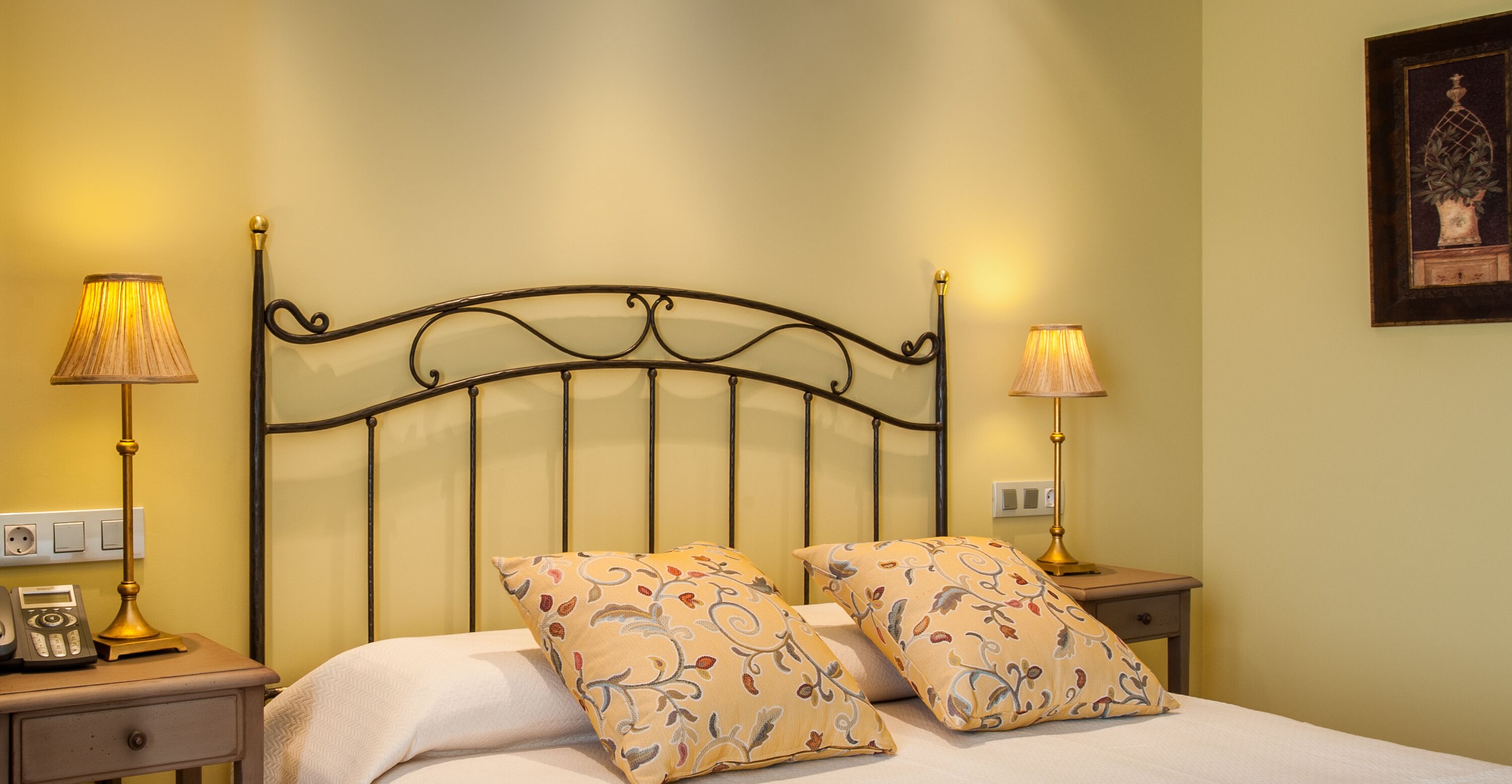 Una cama bien hecha con dos almohadas estampadas, una mesita de noche con una lámpara y un cuadro colgado en la pared.