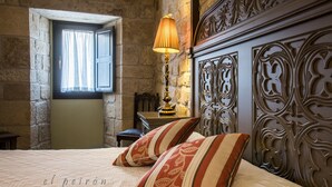 Quarto com parede de pedra, uma cama com duas almofadas decorativas, uma mesa de cabeceira, abajur e janela com cortinas.