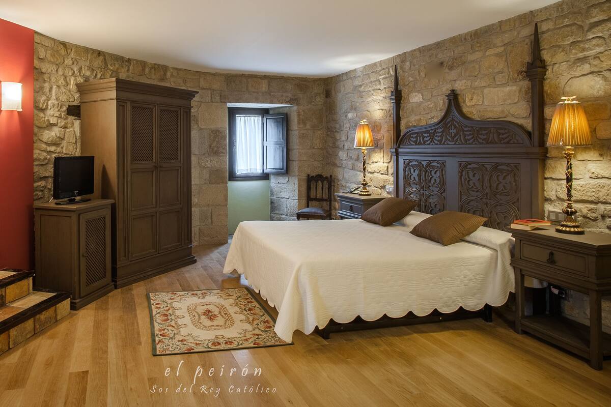 A bedroom with a stone wall, a wooden bed with a headboard, a nightstand, a lamp, a TV on a cabinet, and a staircase.