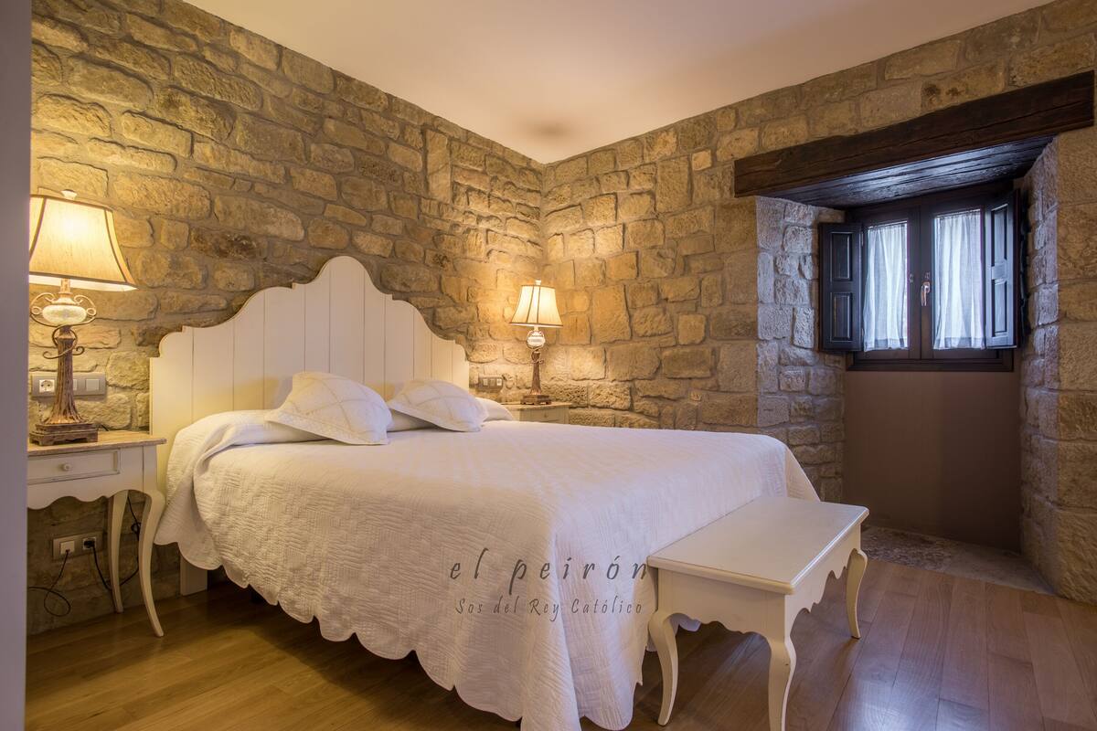 A bedroom with a stone wall, a bed with white bedding, two bedside lamps, and a window with shutters.