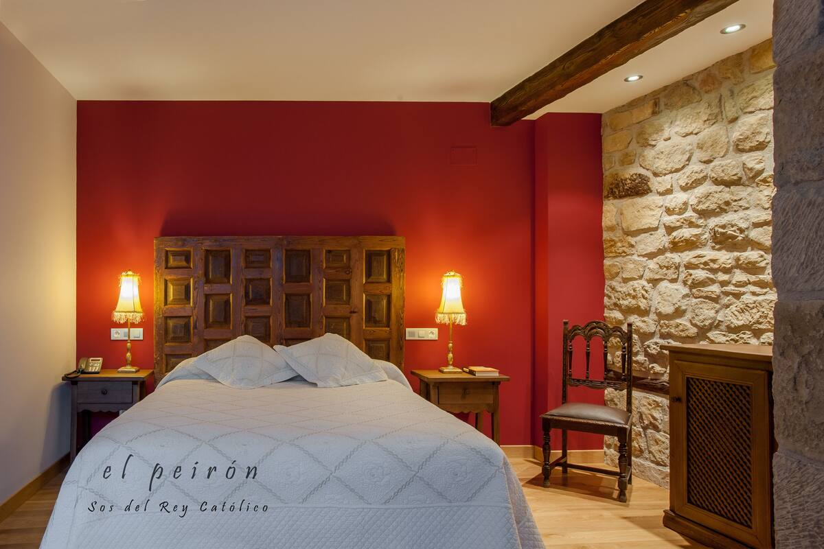 A hotel room with a large bed, two bedside lamps, a wooden chair, and a stone wall.