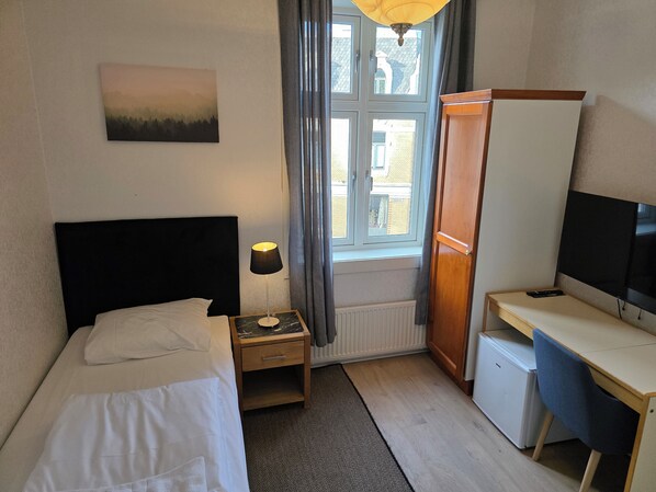 A hotel room with a bed, bedside table, lamp, TV, desk, and a window with curtains. - Grand Hotel Hønefoss (Ringerike)