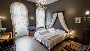 Ein Schlafzimmer mit einem großen Bett, einem Nachttisch, einem Sessel, einer Bank, einer Kristallleuchtre und Vorhängen.