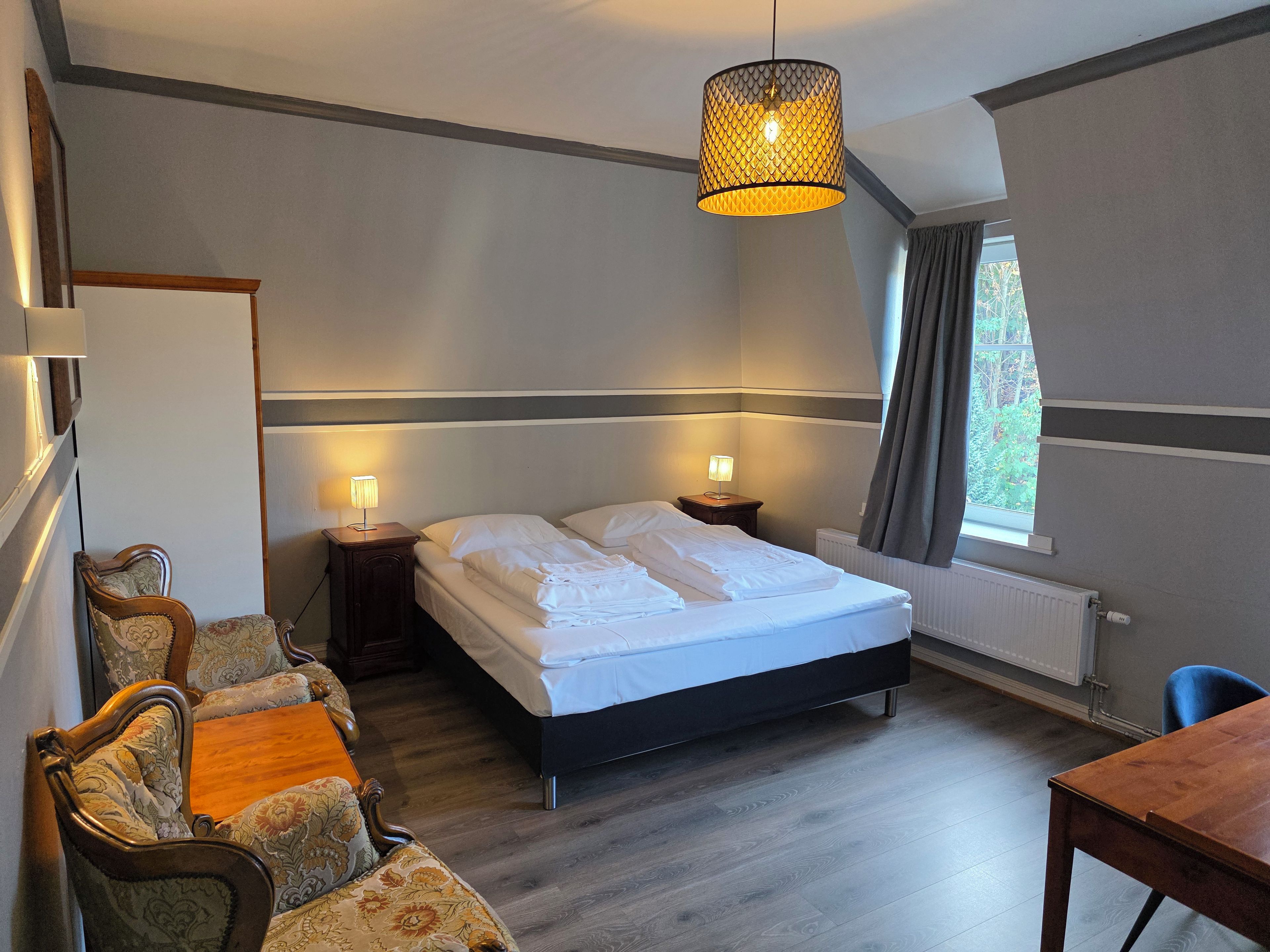 Quarto de hotel com cama, abajures, mesa de madeira, uma cadeira e janela com cortinas.